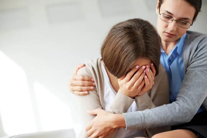 A supportive counselor or friend placing a comforting hand on the shoulder of a woman who is sitting down and crying with her face buried in her hands.
